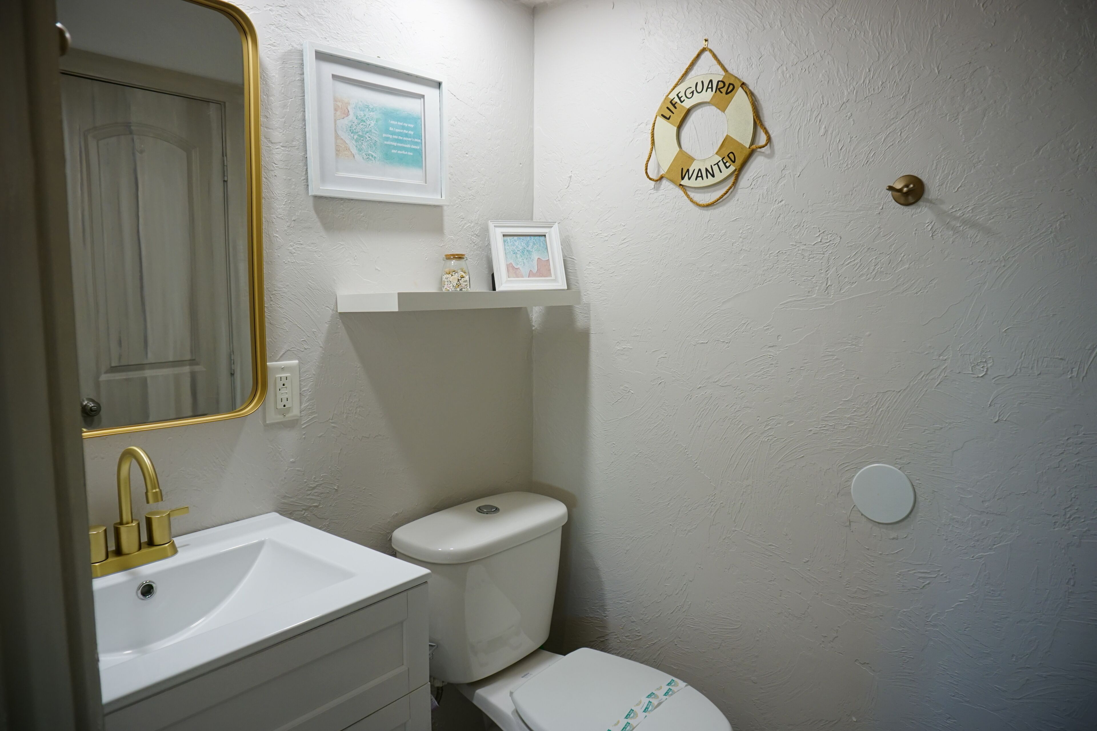 A bathroom with a white sink, gold faucet, and a toilet. There is a framed picture on the wall and a life guard wanted sign.