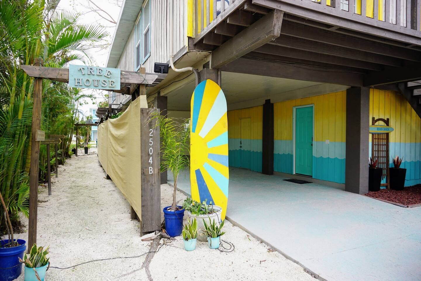A beach house with a colorful surfboard sign, a sandy pathway, and potted plants.