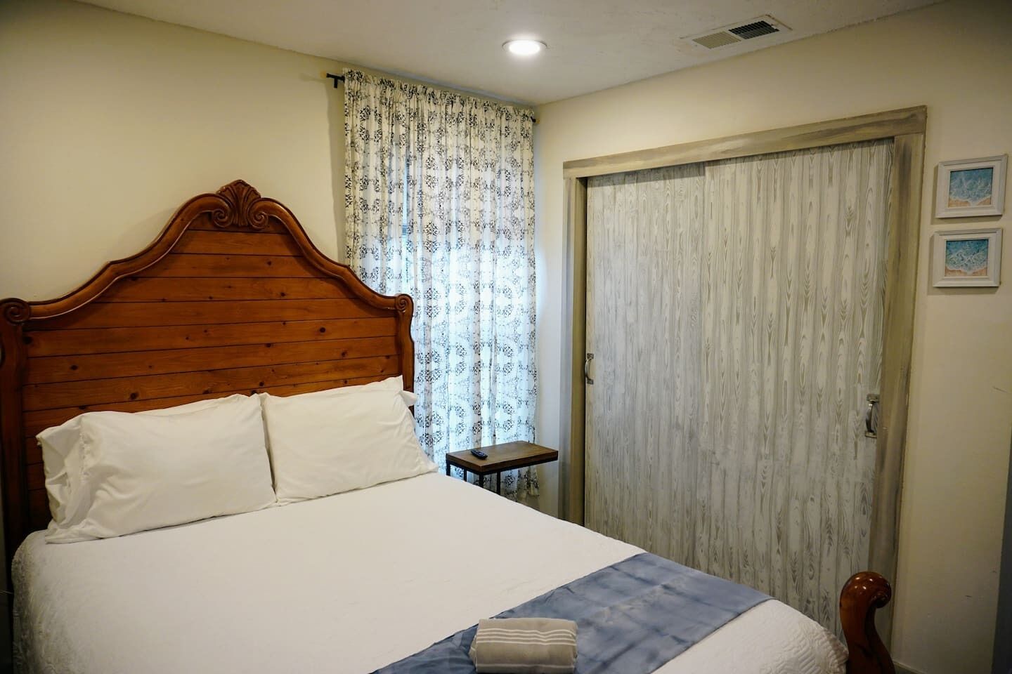 A bedroom with a wooden bed, white bedding, a small bedside table, a curtain, and a door.