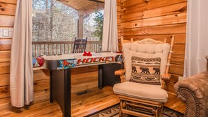 Interior de una cabaña de madera con una mesa con motivos de hockey, un sillón acolchado con diseño de oso y vistas a árboles a través de una ventana.