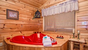 Interior de una cabaña de madera con una bañera de hidromasaje roja, un cuadro en la pared y una ventana con persianas.