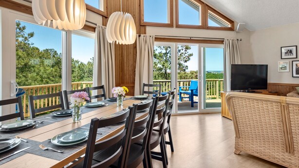 Comedor con una mesa preparada para cuatro, televisor y balcón con vista a árboles.
