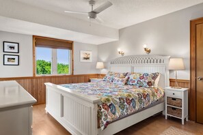 Un dormitorio con cama blanca, cabecera de madera, dos lámparas de noche, ventilador de techo, ventana con vistas y cómoda de madera.