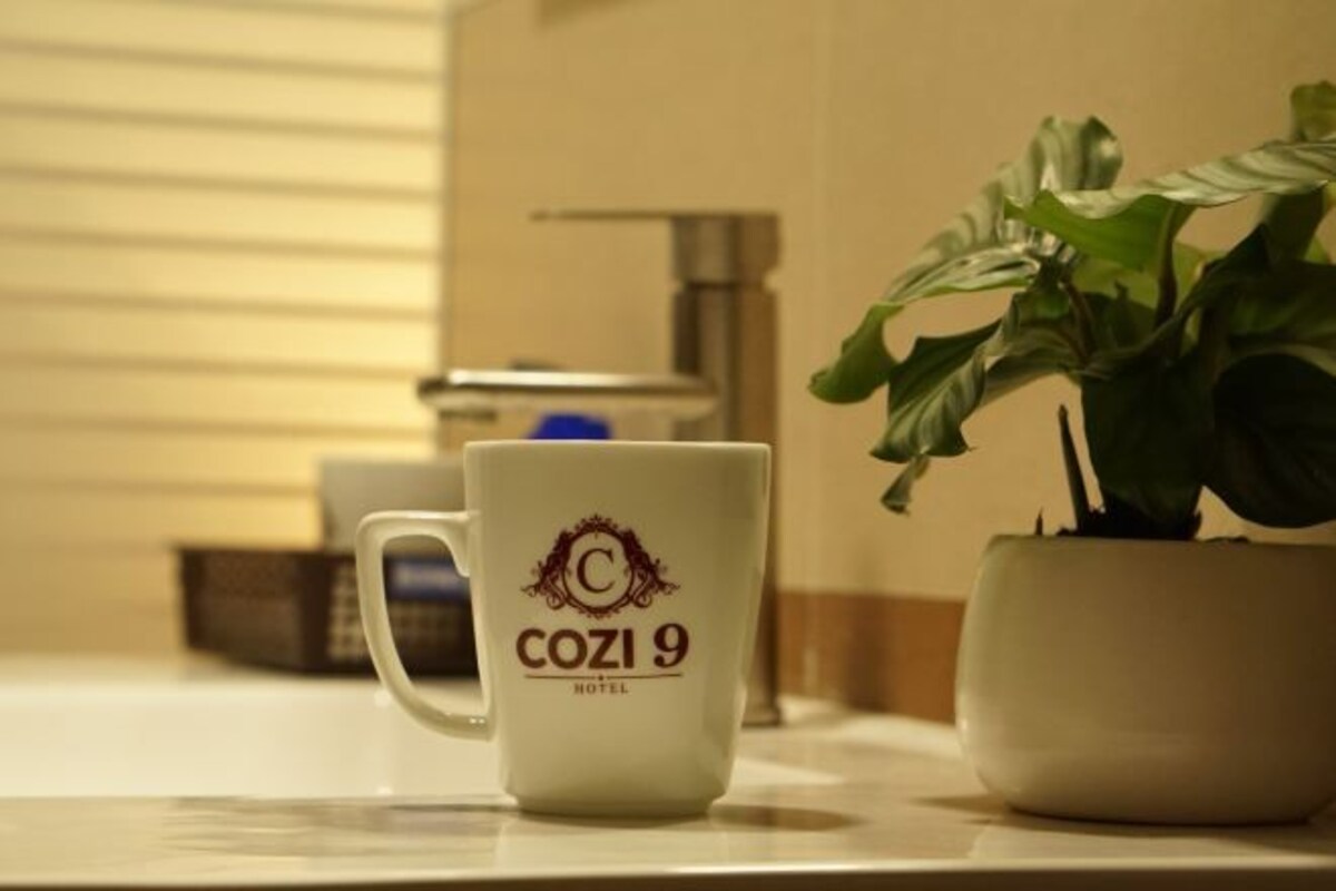 Una taza blanca con el logotipo 'COZI 9 HOTEL' sobre una encimera junto a una planta en maceta.