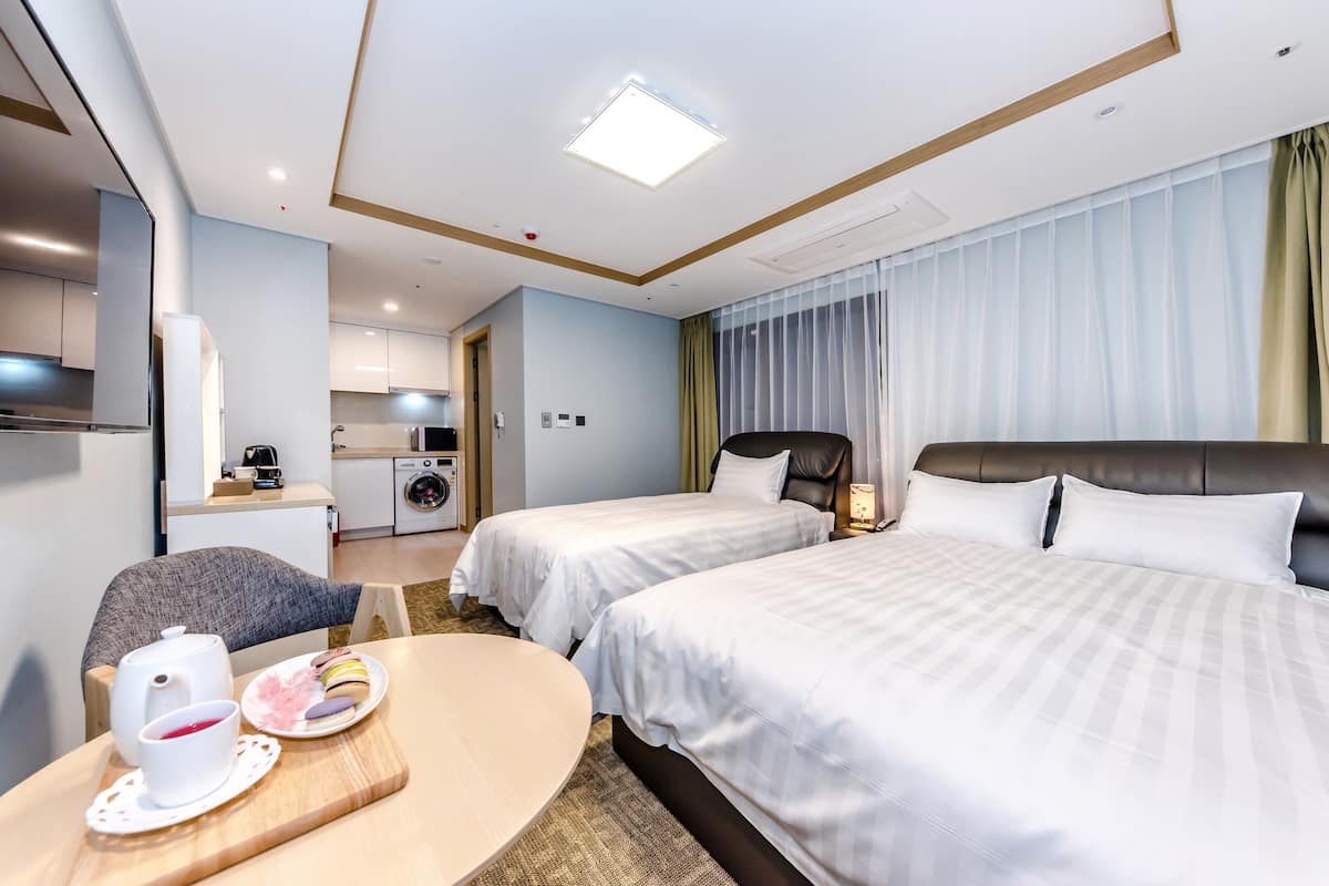 Habitación de hotel con dos camas, una mesa de comedor con tetera y pasteles, una kitchenette y un baño.