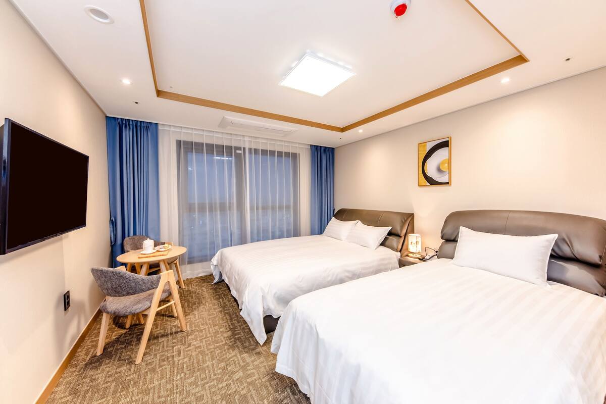 Una habitación de hotel con dos camas, televisión, una mesita con sillas y una ventana con cortinas.