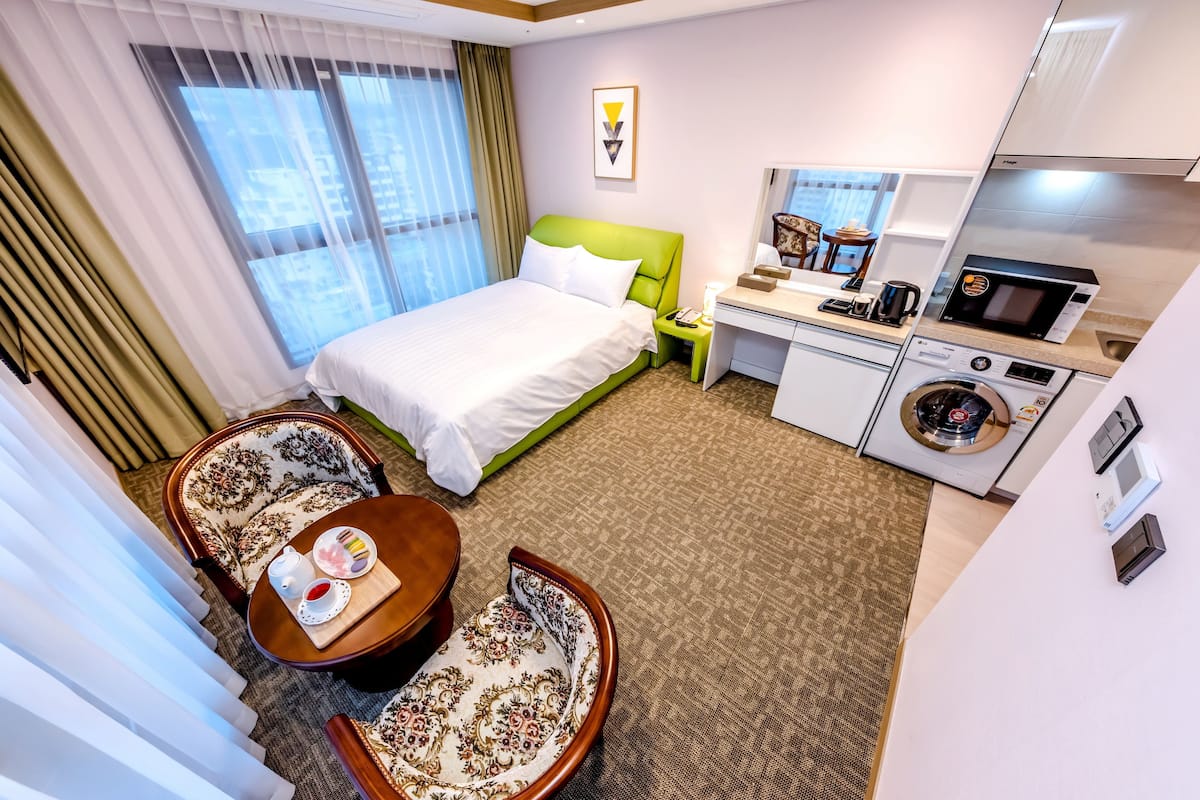 Una habitación de hotel con cama, zona de estar con mesa y sillas, kitchenette con microondas y lavadora, y ventana con cortinas.