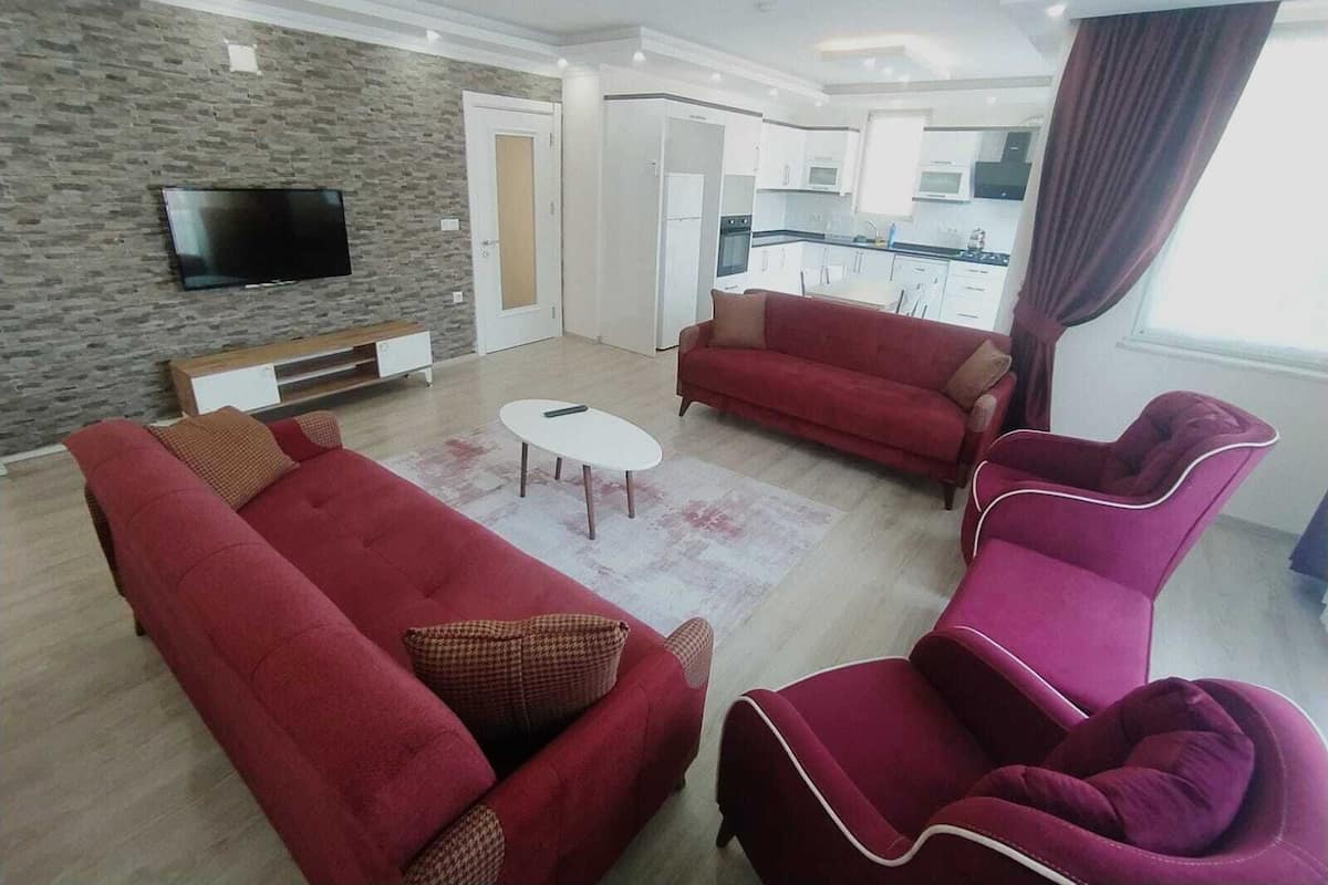 A living room with red sofas, a TV mounted on a stone wall, a kitchen in the background, and a window with curtains.