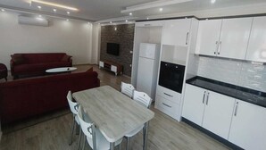 A modern living room with a red sofa, a dining table with chairs, a kitchen with white cabinets and a black countertop, and a TV mounted on a brick wall. - Kaya House Daily Flat (Ortaca)