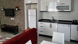 A modern kitchen with white cabinets, a built-in microwave, and a refrigerator. There is a black countertop with a sink and a stove. A flat-screen TV is mounted on a brick wall, and a red sofa is visible in the living area. - Kaya House Daily Flat (Ortaca)