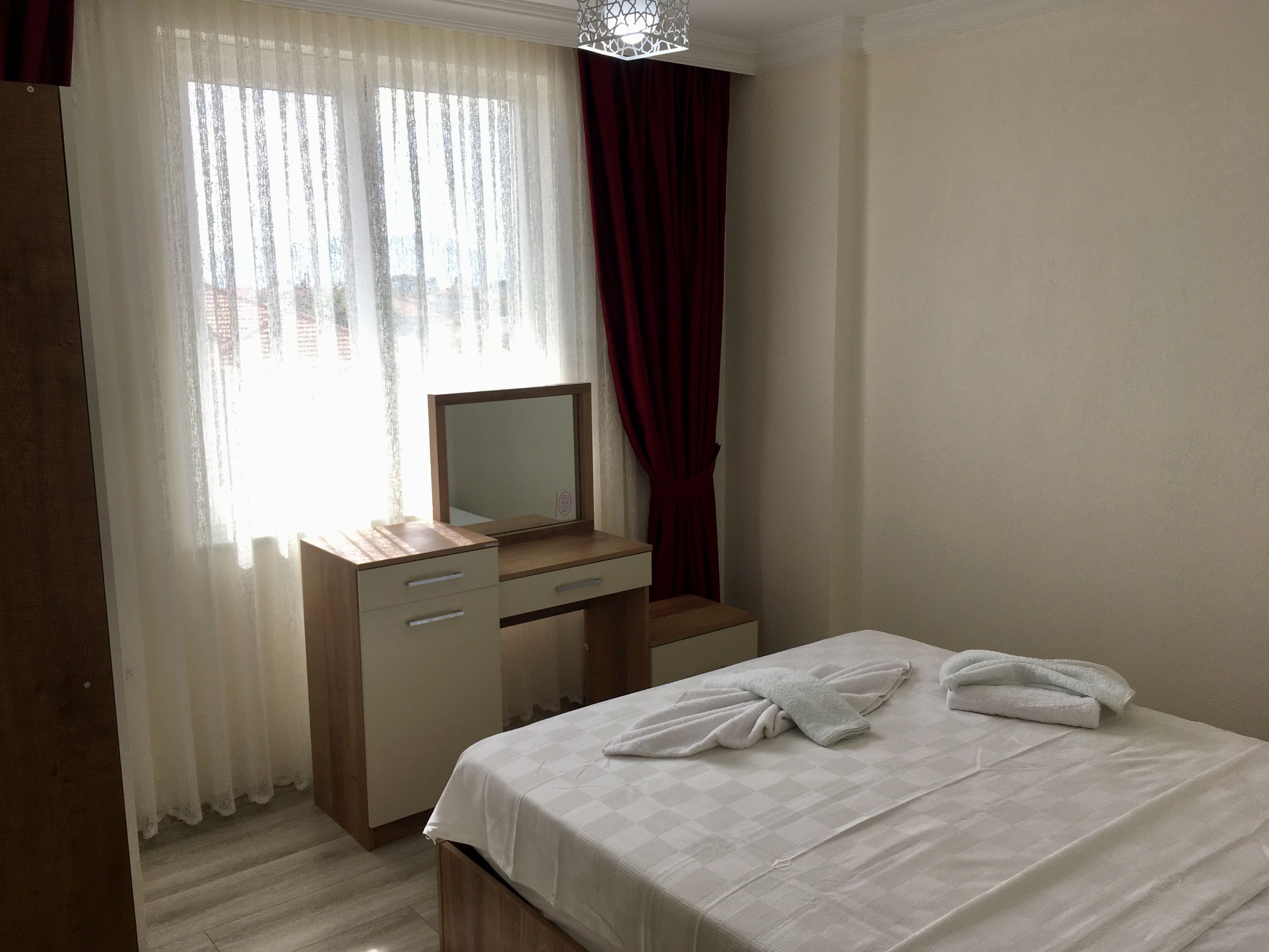 Ein Hotelzimmer mit einem Bett, einem Nachttisch aus Holz mit Spiegel und einem Fenster mit durchsichtigen Vorhängen und einem roten Vorhang.