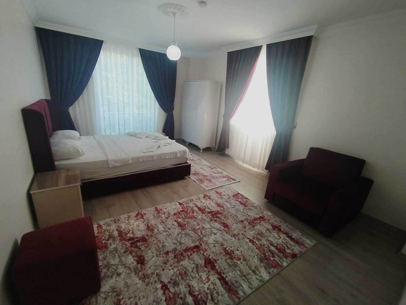A bedroom with a bed, two chairs, a standing heater, and a large window with curtains.