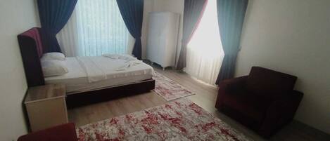 A bedroom with a bed, two chairs, a standing heater, and a large window with curtains.