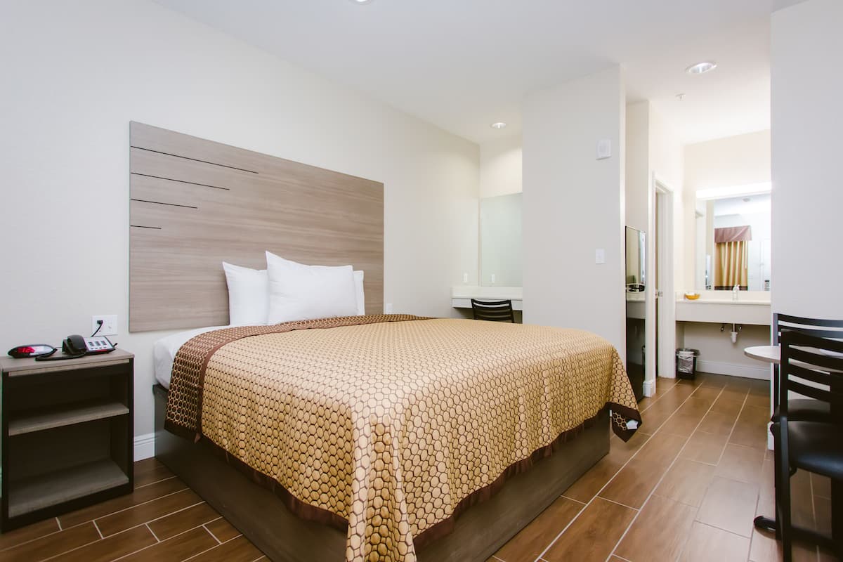 A modern hotel room with a wooden headboard, a large bed with a patterned bedspread, a bedside table with a phone, and a small shelf.