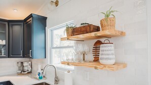 A kitchen with dark cabinets, a white countertop, a sink, and wooden shelves holding plants and glassware. - ★ "The house is perfection—stylish, comfortable + close to the beach." (Nags Head)