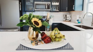 A kitchen counter with a white plate holding a sunflower, a bottle, a bunch of bananas, and some apples. - ★ "The house is perfection—stylish, comfortable + close to the beach." (Nags Head)