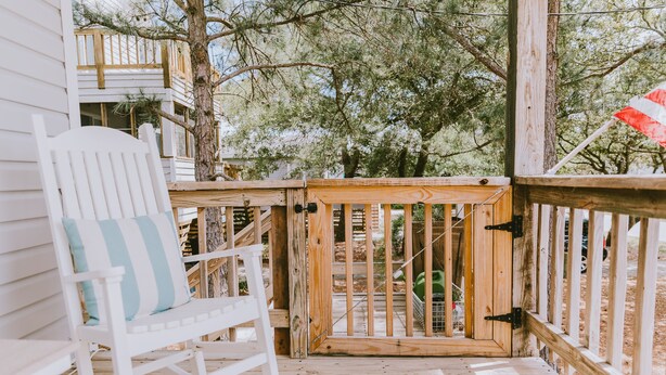 A wooden deck with a white rocking chair and a gate leading to a garden area. - ★ "The house is perfection—stylish, comfortable + close to the beach." (Nags Head)