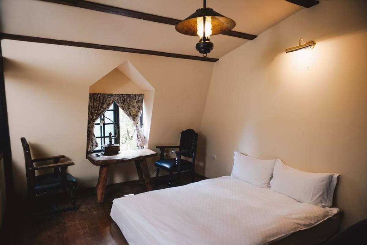 A bedroom with a large bed, a desk with a chair, a window with curtains, and a hanging light fixture.