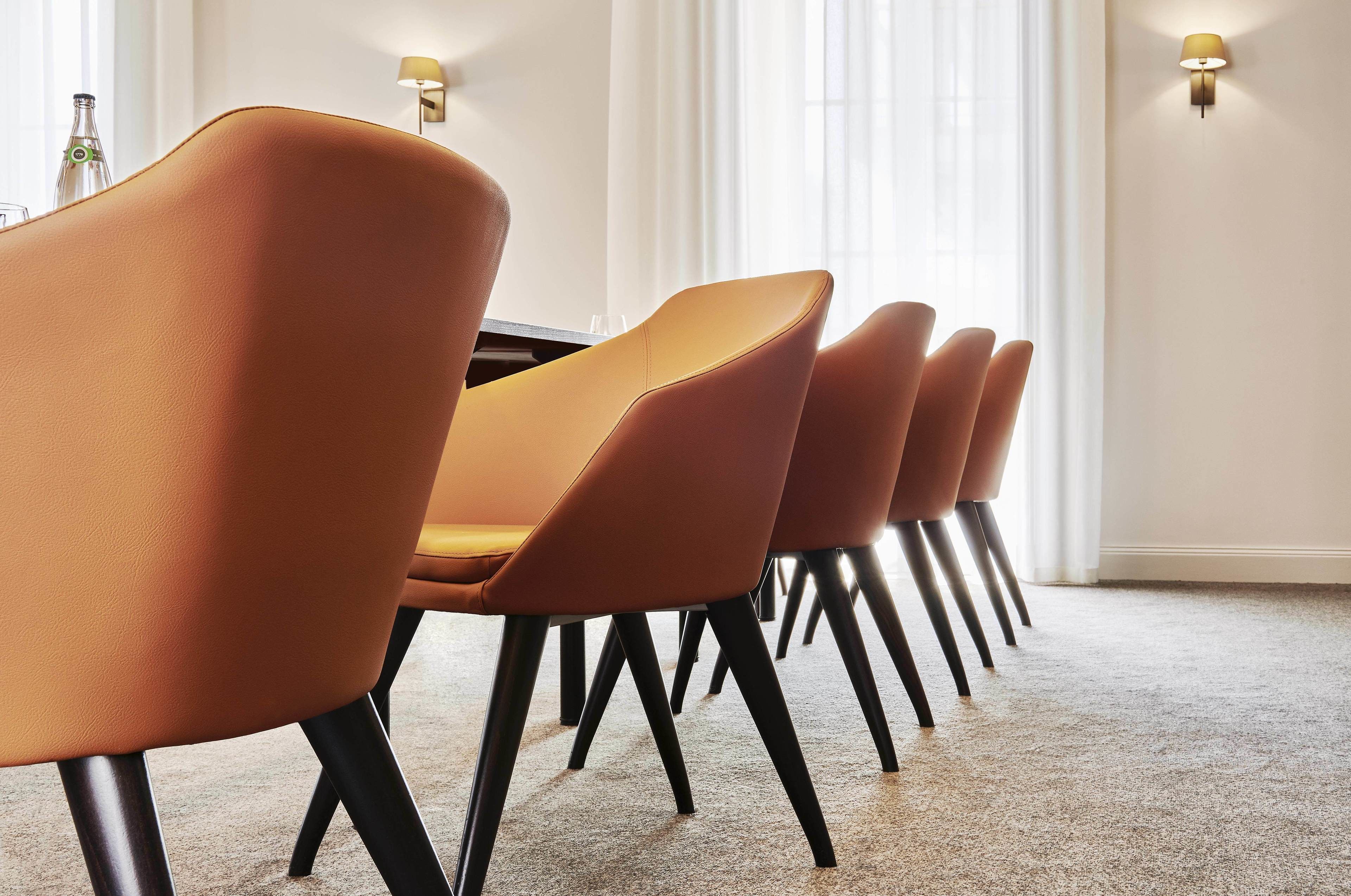 A row of modern orange chairs with black legs, arranged in a dining area with a carpeted floor and a table.