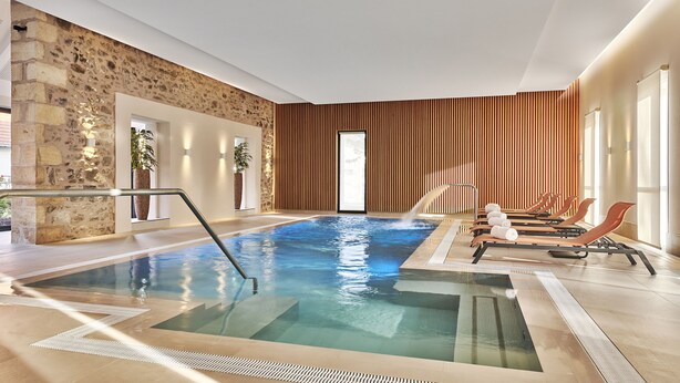 A modern indoor pool with clear water, wooden loungers, and a stone wall. - Château Saint-Jean (Montlucon)