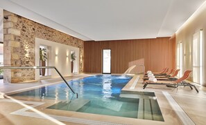 A modern indoor pool with clear water, wooden loungers, and a stone wall. - Château Saint-Jean (Montlucon)