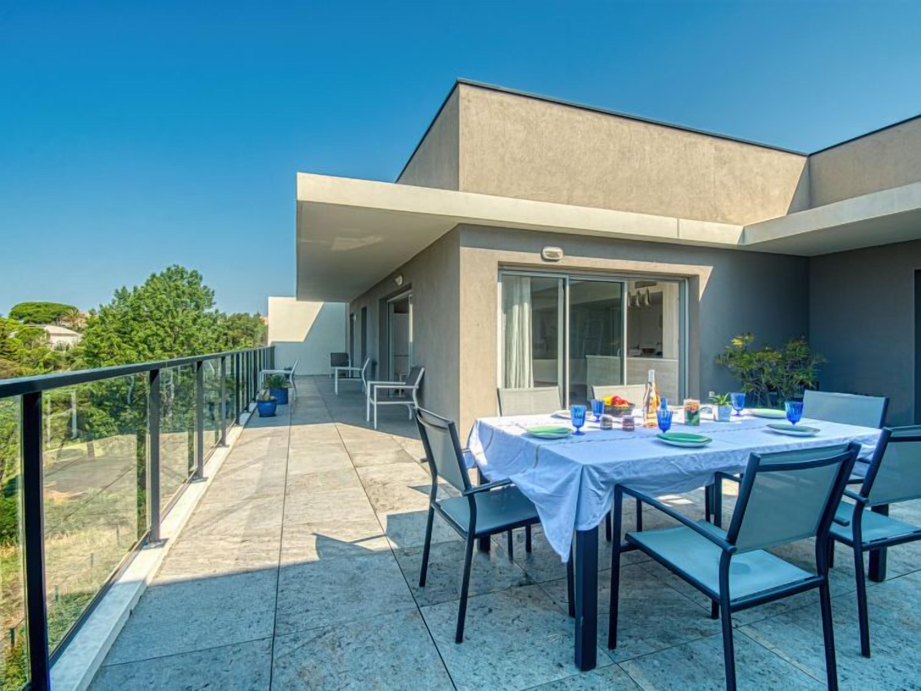 Un espace repas extérieur moderne, avec une table dressée pour quatre personnes, entourée de chaises, sur un balcon offrant une vue sur la verdure et un bâtiment en arrière-plan.