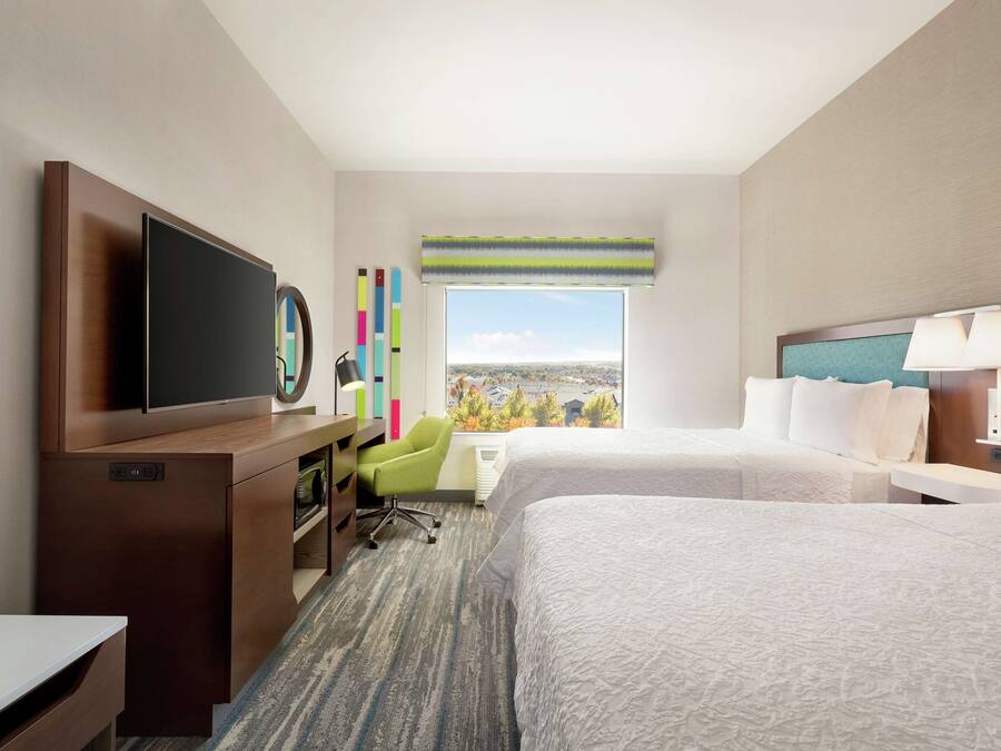Quarto de hotel com duas camas, uma escrivaninha com cadeira, televisão e janela com vista para a cidade.