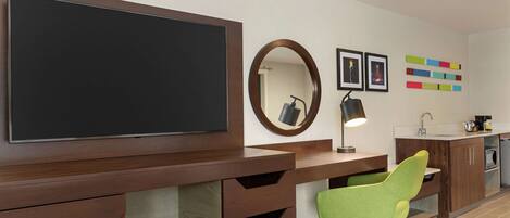 A modern kitchen with a large flat-screen TV, a green chair, wooden cabinets, a sink, and a mirror.