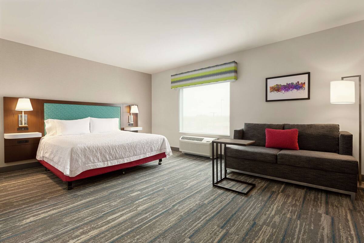 Une chambre d’hôtel comprenant un lit, un canapé, une fenêtre avec des stores, un climatiseur fixé au mur et un tableau encadré accroché au mur.