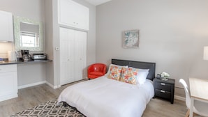 A bedroom with a bed, a nightstand, a chair, a desk, a red chair, a coffee maker, a mirror, and a door.