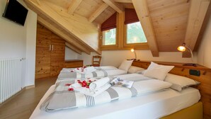 A bedroom with a sloped ceiling, wooden beams, a bed with white bedding and rose petals, a bedside table with a lamp, and a TV on the wall.