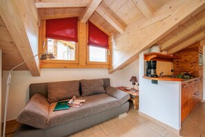 Quarto aconchegante com teto de madeira, um sofá, uma mesa pequena e uma cozinha compacta.