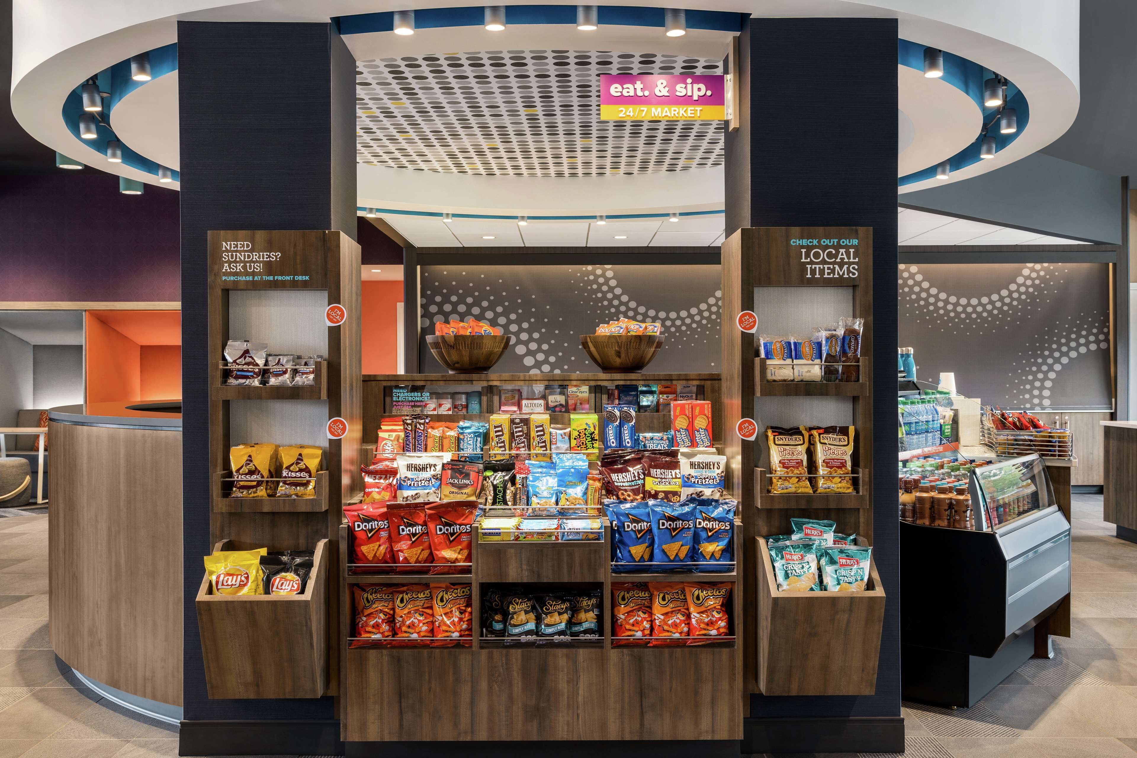 A snack kiosk with a variety of chips and snacks, a 24/7 market sign, and a 'need sundries?' sign.