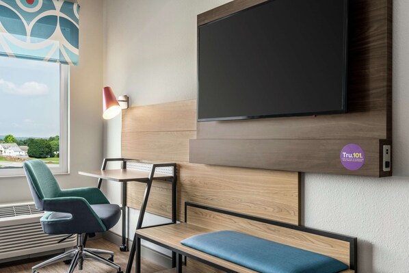 Habitación moderna con una televisión de pantalla plana grande montada en la pared, un escritorio con una silla, un banco y una ventana con vistas a zonas verdes y edificios.