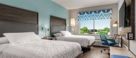 A hotel room with two beds, a desk, a chair, a TV, and a window with a view of greenery.