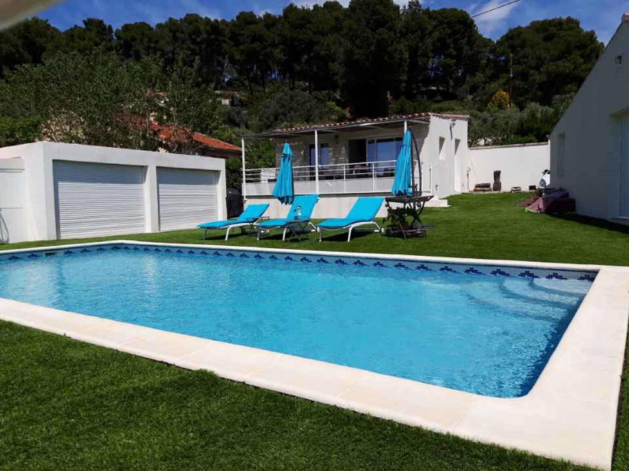 Une piscine rectangulaire bordée de carreaux, entourée d’une pelouse bien entretenue. Quatre chaises longues bleues sont alignées le long du bord de la piscine.