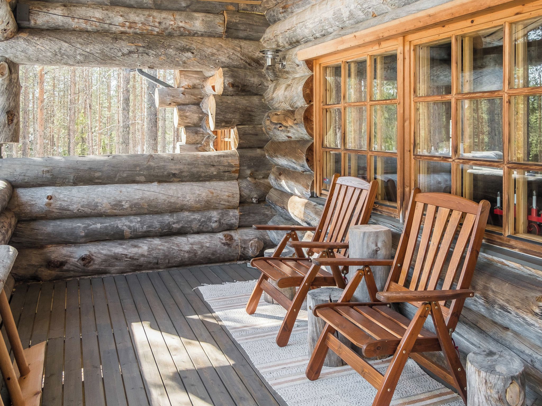 A log cabin interior with a wooden deck, two Adirondack chairs, and a large window with multiple panes.