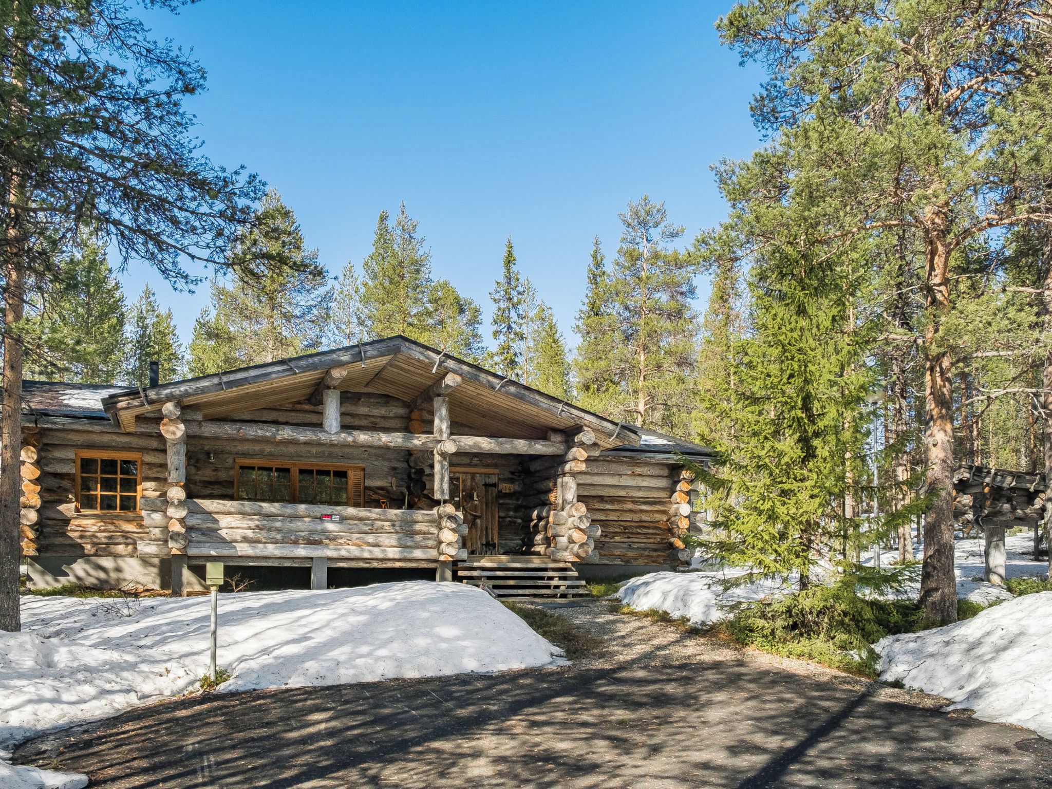 Una cabaña de troncos con porche y chimenea, rodeada de nieve y árboles.
