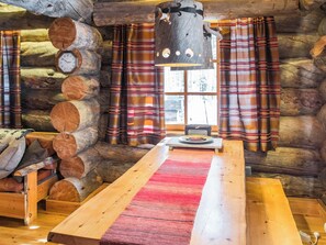 Interior de uma cabana de tronco com uma mesa de madeira, cortinas xadrez e um relógio na parede.