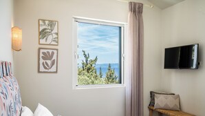 A bedroom with a large window, a bed, a bedside table with a TV, and wall art. - Villas Muscallas Elia by Konnect - 200m from the beach (Paxoi)