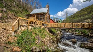 Eine rustikale Holzbrücke führt zu einem Steinhaus mit einem Metall-Dach, mit Blick auf einen Fluss.