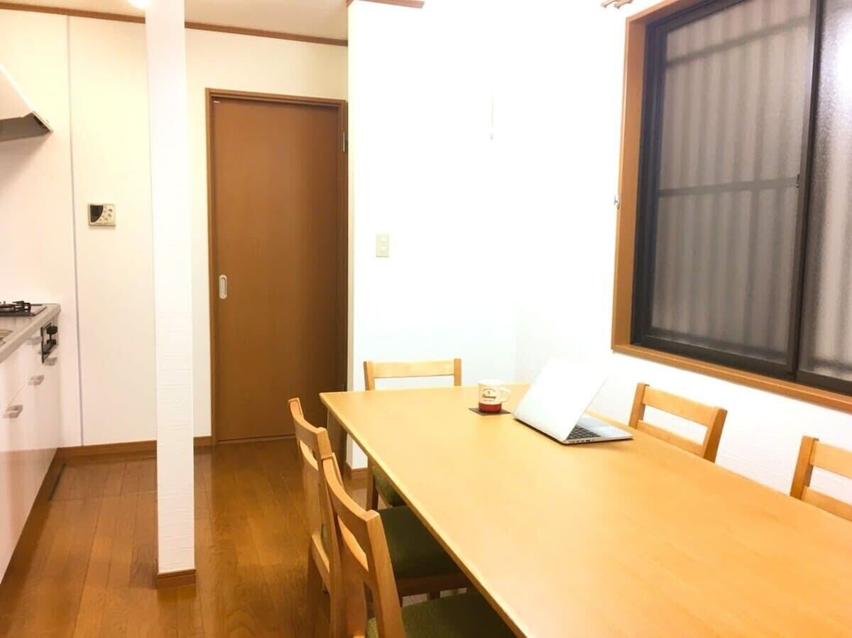 A room with a wooden dining table, chairs, a laptop, and a window with blinds.