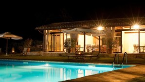 A swimming pool with lounge chairs and umbrellas, a covered patio area with tables and chairs, and a well-lit building in the background. - Le Case Residenza di Campagna (Assisi)