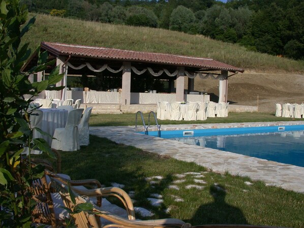 Zona de piscina con un área de estar cubierta y mesas al aire libre, rodeada de vegetación.