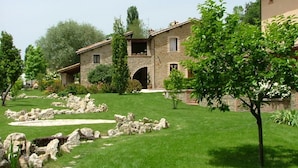 A stone building with a sloped roof, a stone archway entrance, and a well-maintained lawn with scattered rocks. - Le Case Residenza di Campagna (Assisi)
