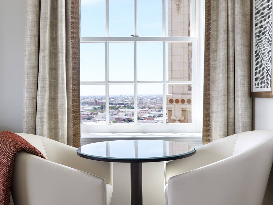 Um quarto com uma mesa redonda, duas cadeiras brancas, vista para a cidade através de uma janela ampla e um painel de parede estampado.