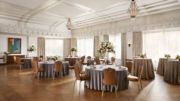A spacious banquet hall with round tables set for dining, elegant chandeliers, and large windows with curtains.