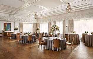 A spacious banquet hall with round tables set for dining, elegant chandeliers, and large windows with curtains.