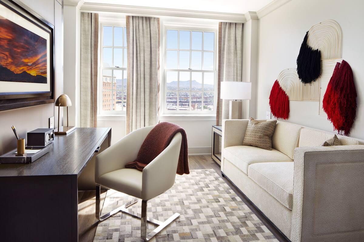A modern hotel room with a desk, chair, sofa, and large windows offering a city view.