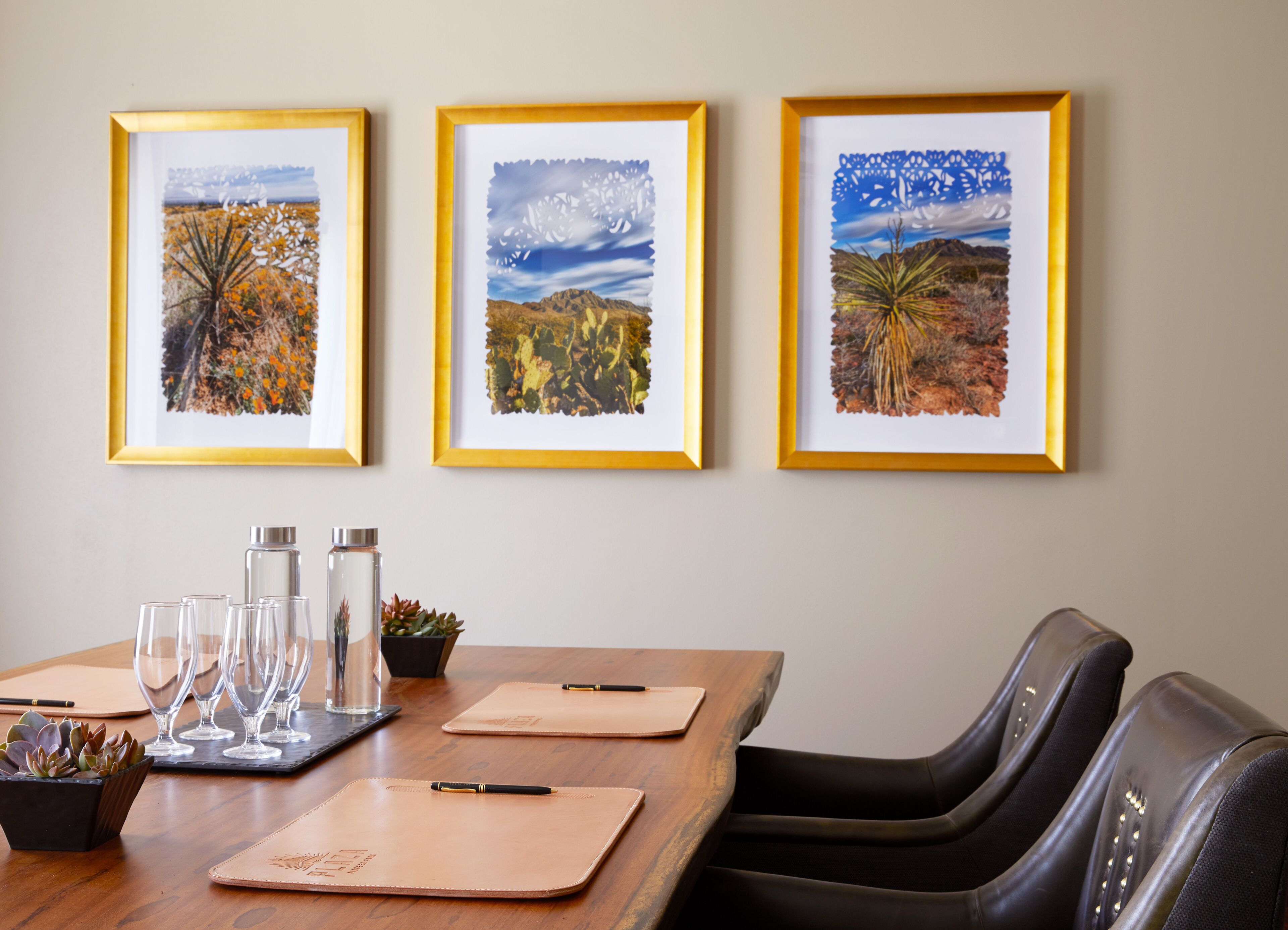 Un espace repas comprenant une table en bois, trois tableaux représentant des paysages encadrés et un ensemble de chaises.